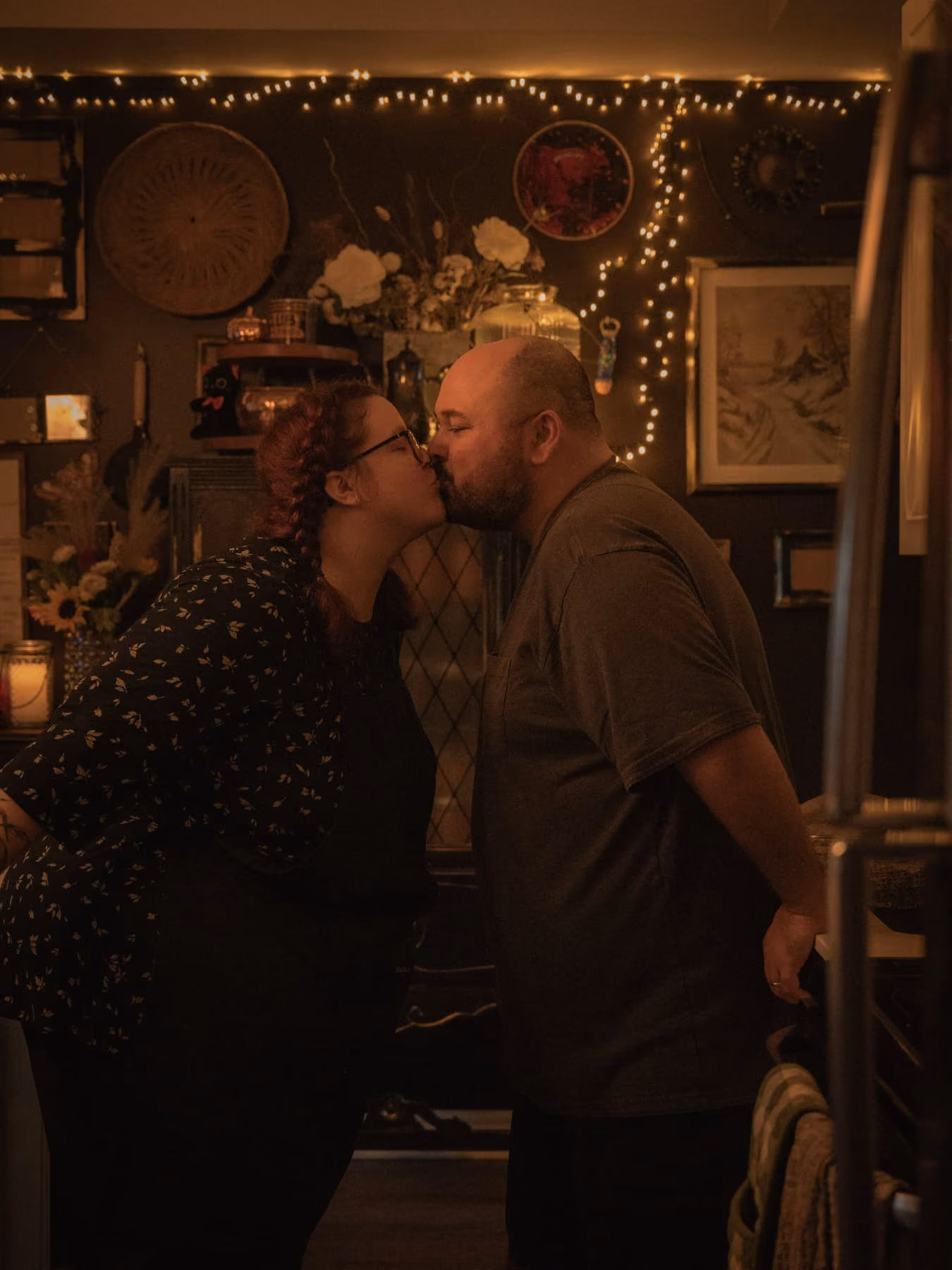 A couple kiss in their kitchen under warm string lights.