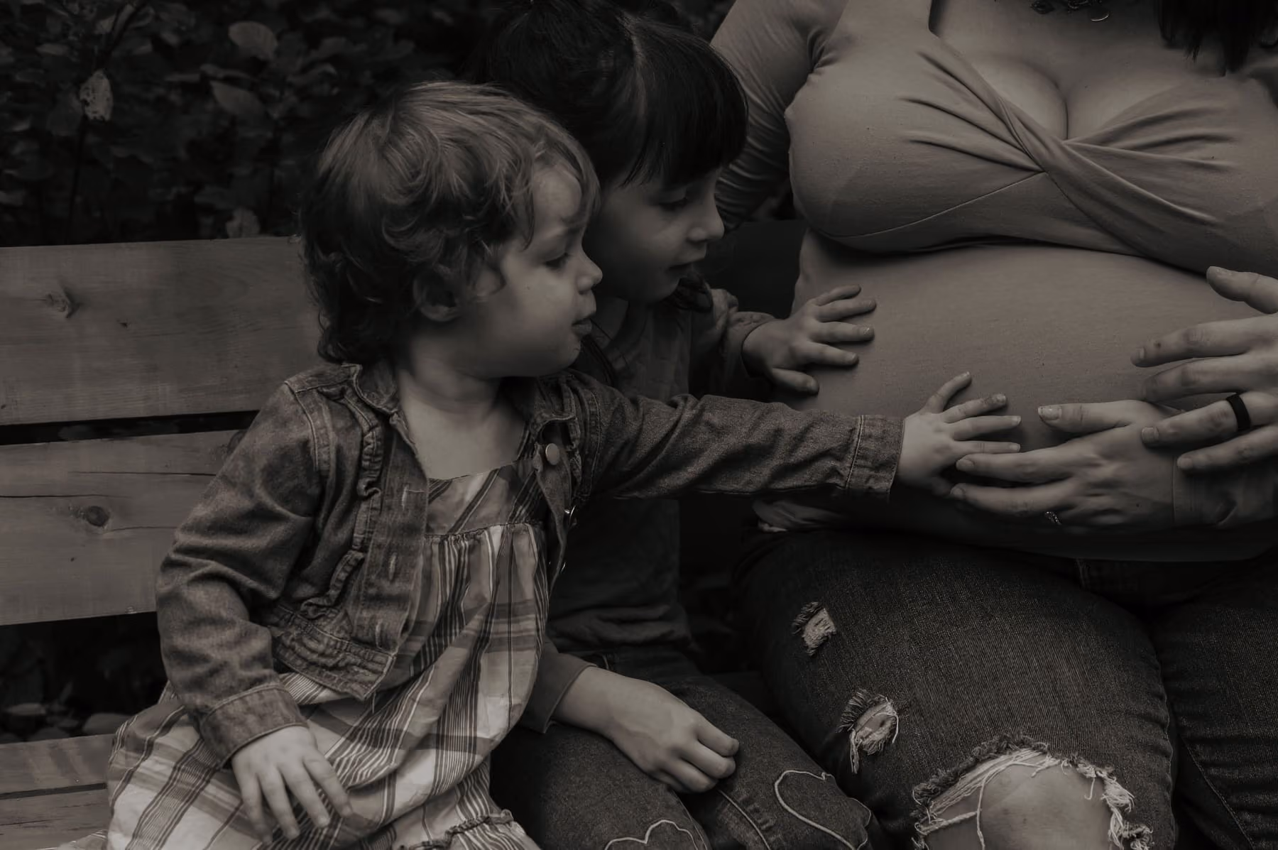 In black and white, two small children reach for their mother’s belly.
