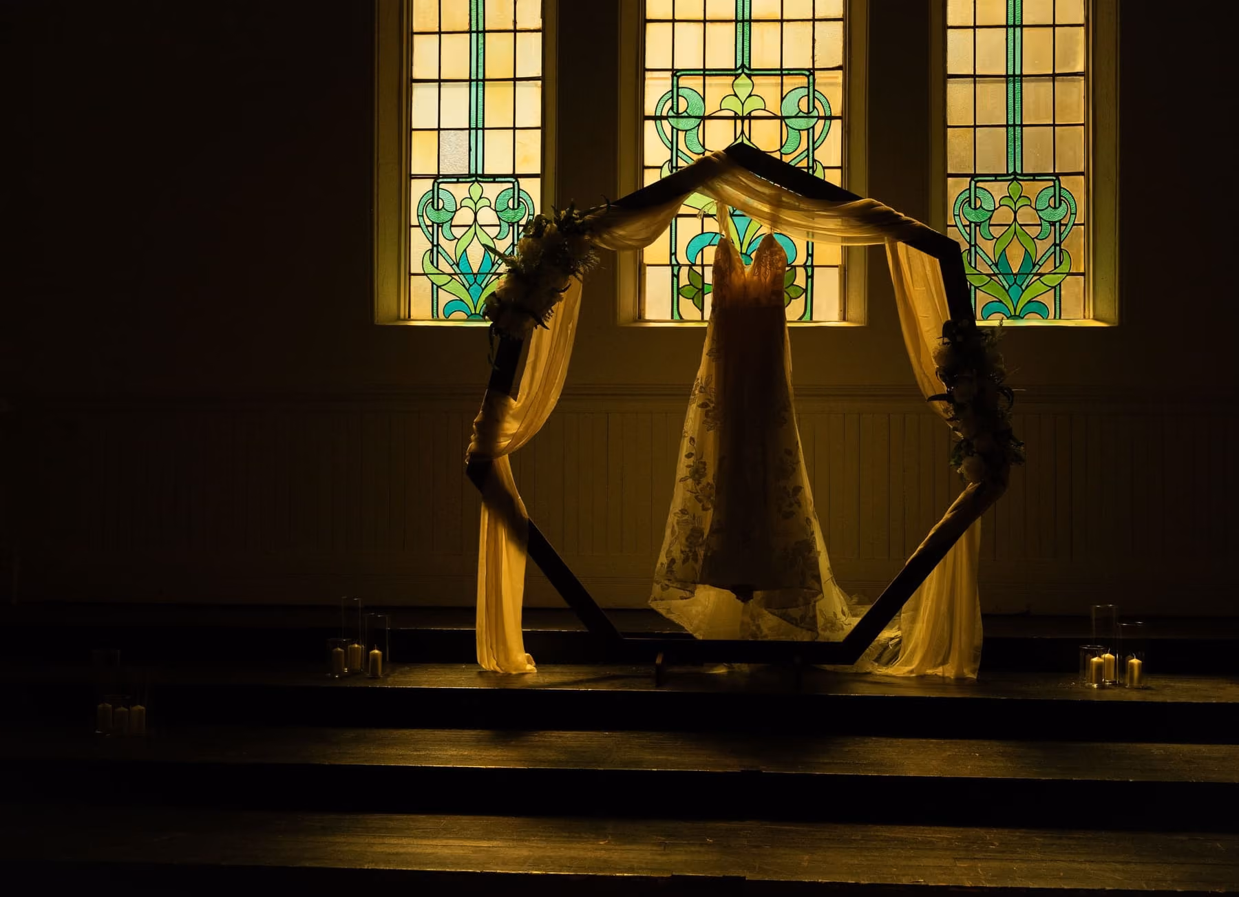 The wedding gown hangs in a hexagon arch, lit gold against stained glass windows.