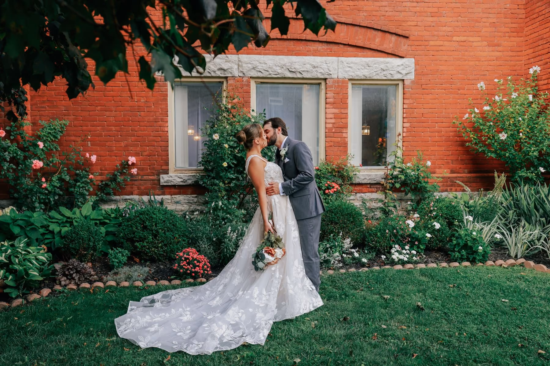 The couple kiss in a garden beside a red brick wall, her gown spread on the grass.